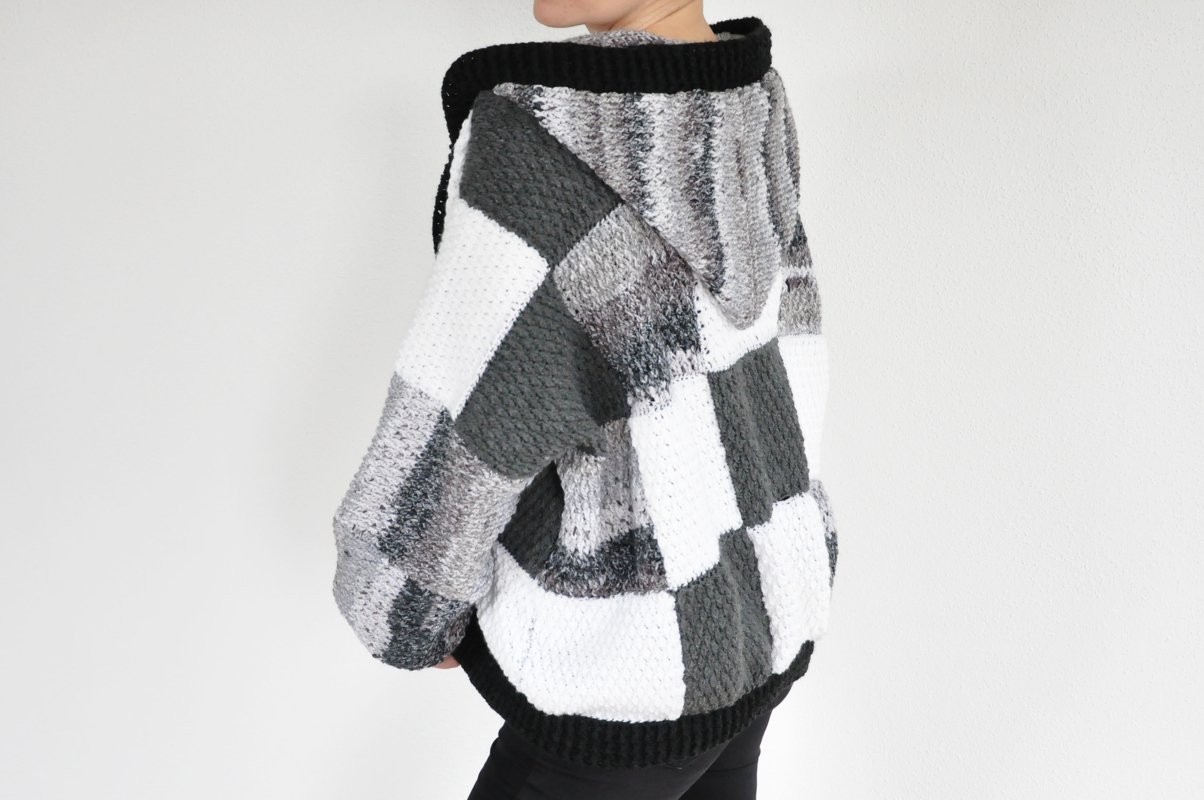 Back view of a person wearing an oversized hooded crochet jacket with a grayscale patchwork of white, gray, and black blocks.