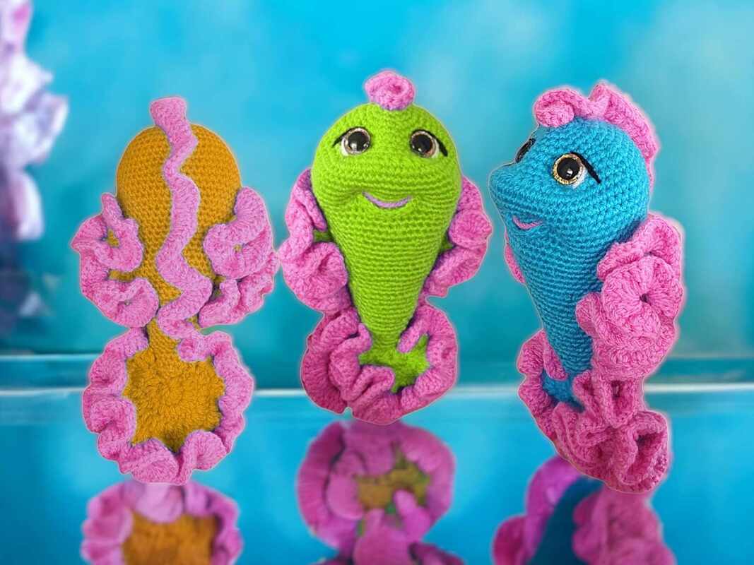 Drei gehäkelte Amigurumi-Wasserbewohner in Gelb, Grün und Blau mit pinken Rüschen auf spiegelnder Fläche