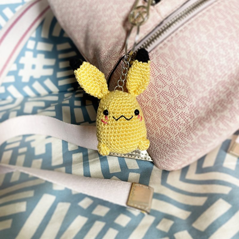 Yellow crocheted amigurumi character keychain with black-tipped ears, wavy embroidered smile, and rosy cheeks attached to a pink bag.