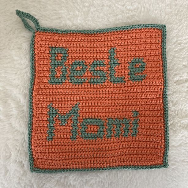 Gehäkelter quadratischer Topflappen in Orange mit grünblauer Schrift 'Beste Mami' und Aufhängeschlaufe