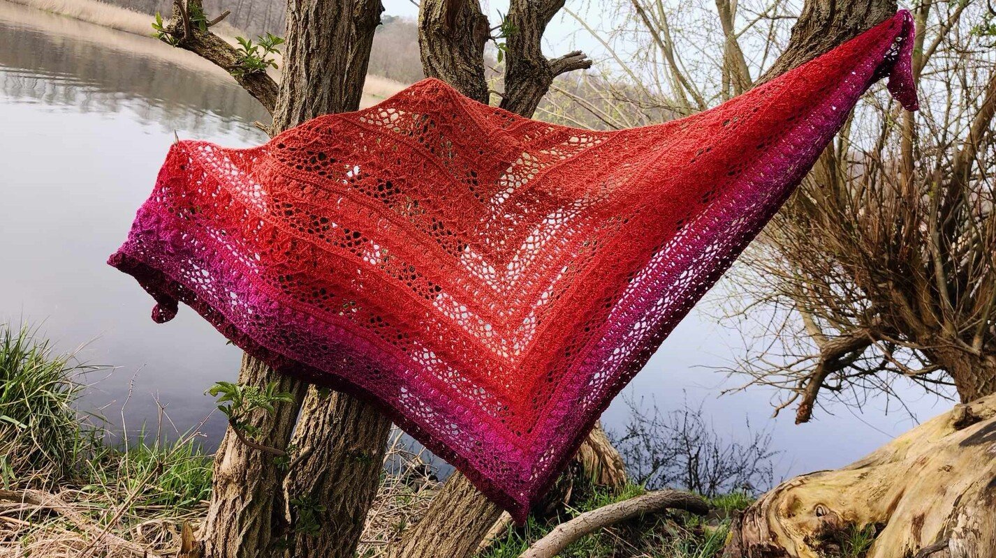 Gehäkeltes Dreieckstuch in Rot‑ und Pinktönen mit Ajourmuster, über einem Baum am Ufer drapiert