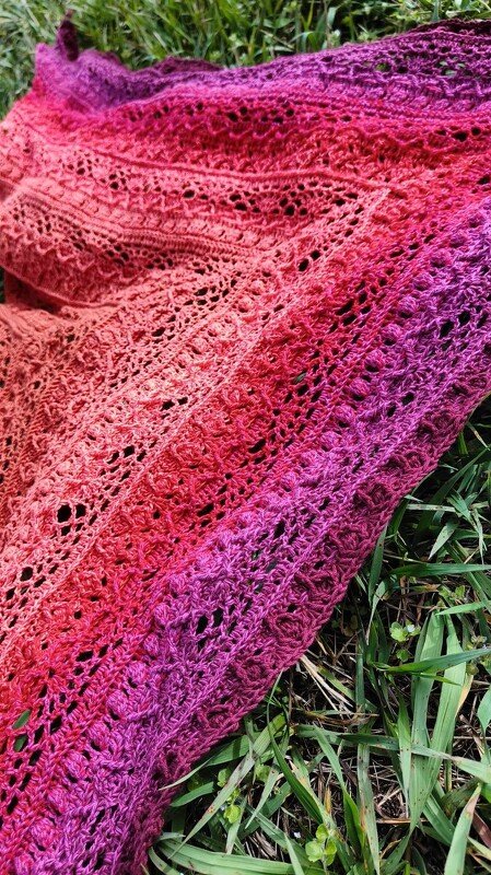 Gehäkeltes Dreieckstuch mit Ajourmuster in Rosa- und Purpurtönen auf Gras