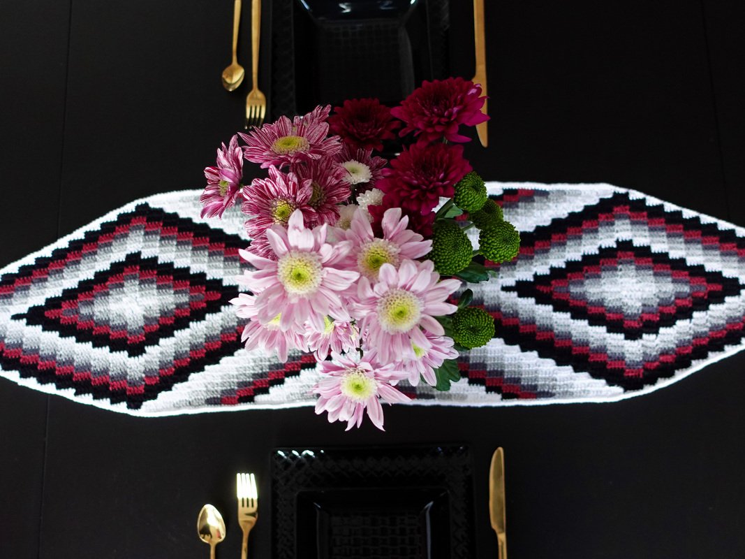 Top view of a crocheted diamond-pattern table runner with pink and burgundy flowers and gold cutlery on a black table