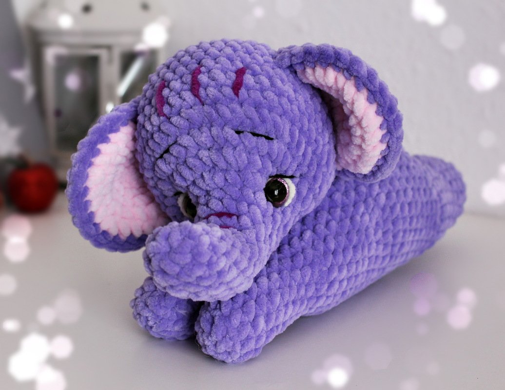 Lila gehäkelter Amigurumi-Elefant mit rosa Ohreninnenflächen, auf heller Fläche liegend.