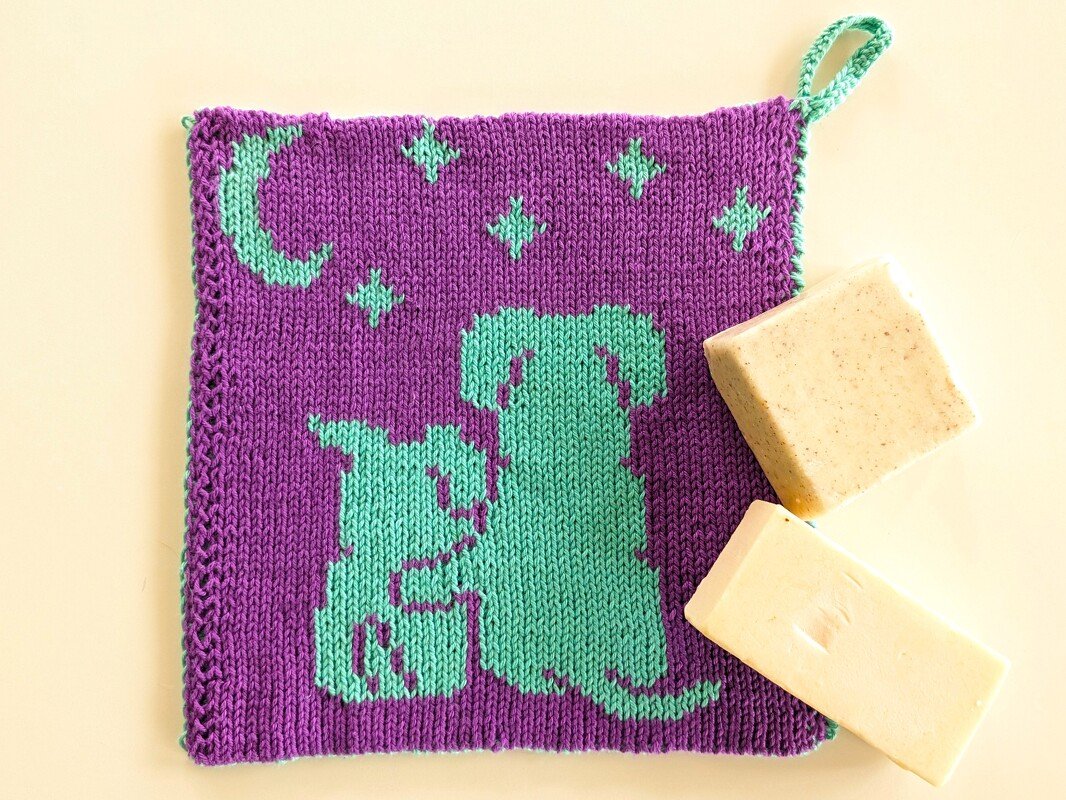 Violetter, gestrickter Topflappen mit mintgr
ner Silhouette von Hund und Katze, Mond und Sternen sowie Aufh
ngeschlaufe; zwei Seifenst
cke daneben