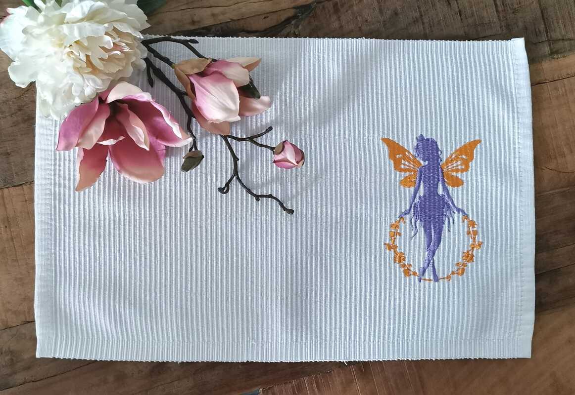 Gestickte violette Elfe mit orangefarbenen Flügeln und Blumenkranz auf weißem, geripptem Stoff, daneben rosa-weiße Blüten
