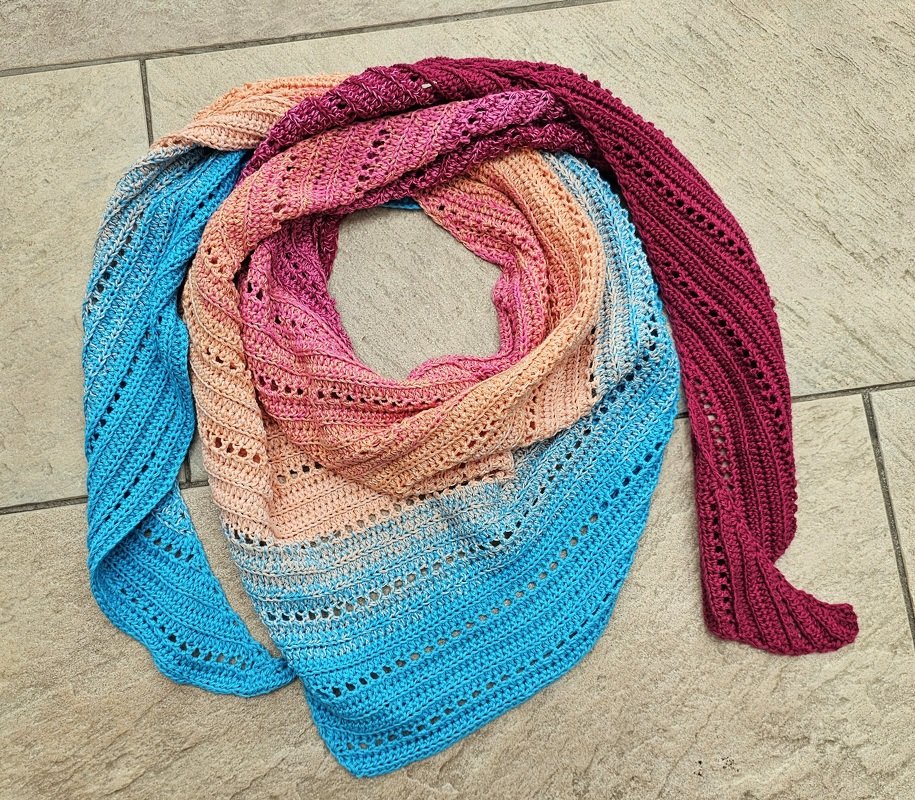 Gehäkeltes Dreieckstuch mit Farbverlauf in Blau, Orange, Pink und Magenta auf hellem Fliesenboden.