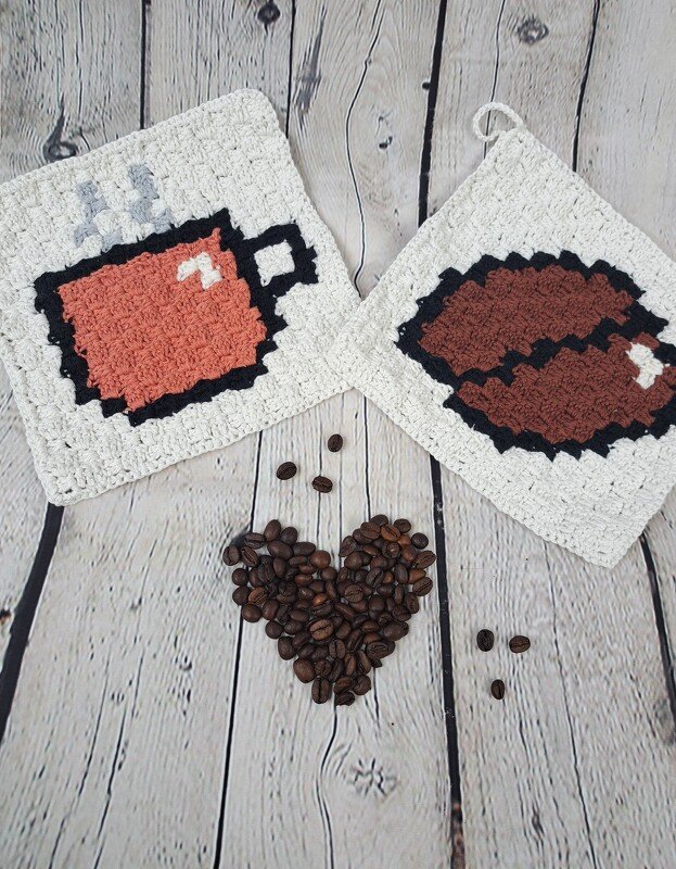 Zwei gehäkelte quadratische Topflappen mit einer Kaffeetasse und einer Kaffeebohne im Pixel‑Design auf hellem Holz, dazu Kaffeebohnen in Herzform.