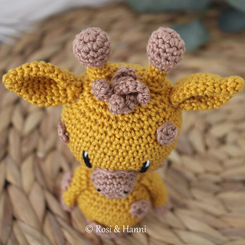 Nahaufnahme einer gehäkelten gelben Amigurumi-Giraffe mit hellbraunen Details auf einer geflochtenen Matte.