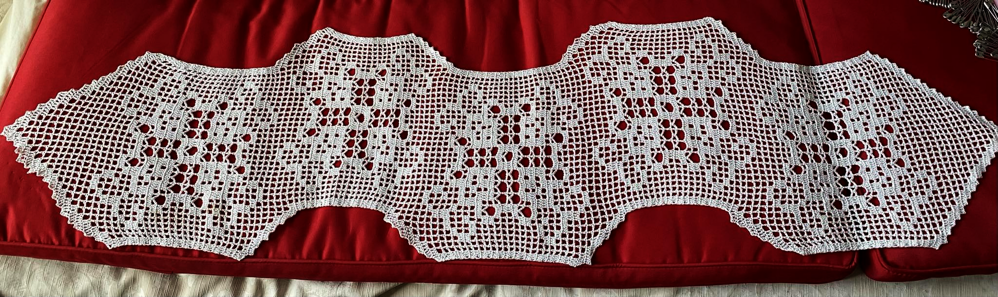 White filet crochet table runner with scalloped wavy edges on a red background.