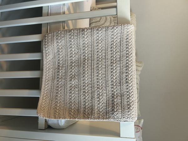 Beige crocheted baby blanket with textured stitch draped over white crib rail