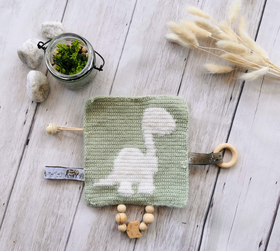 Green crocheted square baby toy with white dinosaur appliqué, wooden teething ring, wooden beads, a small bell, and a patterned fabric tag.