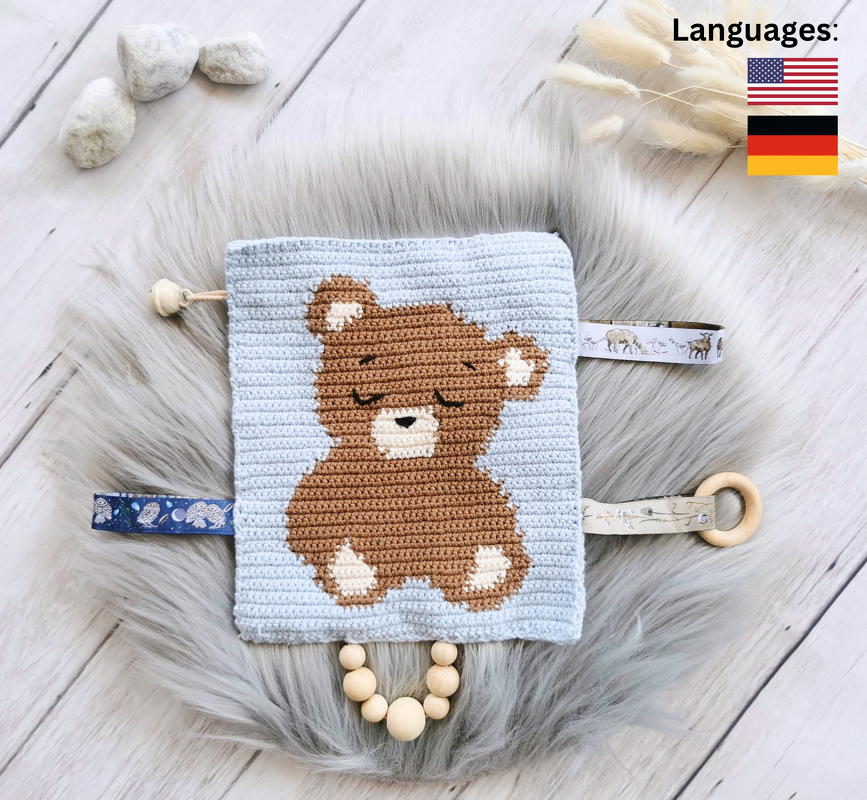 Flat-lay of a fast-to-crochet baby tag blanket with central teddy pattern, ribbon tags and wooden bead loop