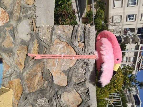 Pink crocheted flamingo amigurumi with yellow beak and black eye resting on a stone wall outdoors.