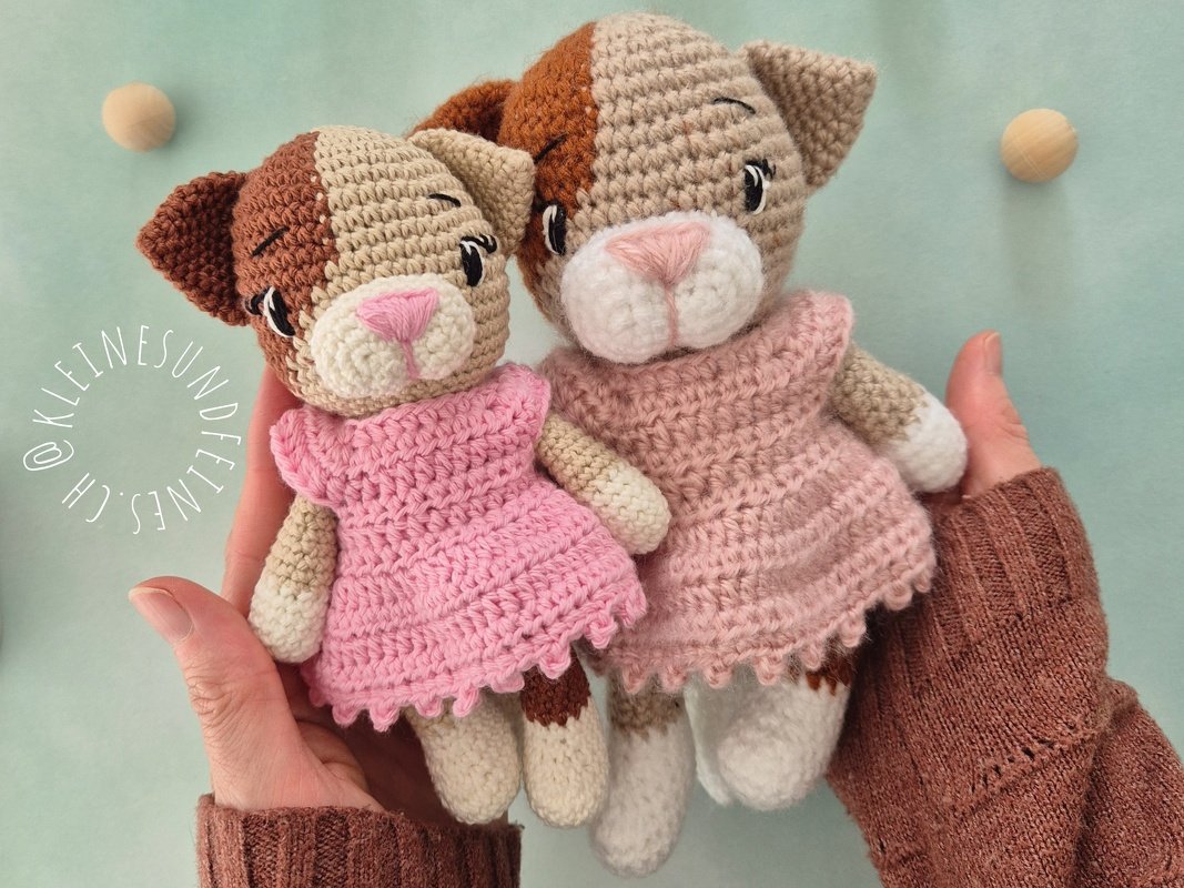 Zwei handgehäkelte Amigurumi-Katzen mit braun-beigem Fell, weißen Pfoten und rosa Kleidern, in Händen gehalten.