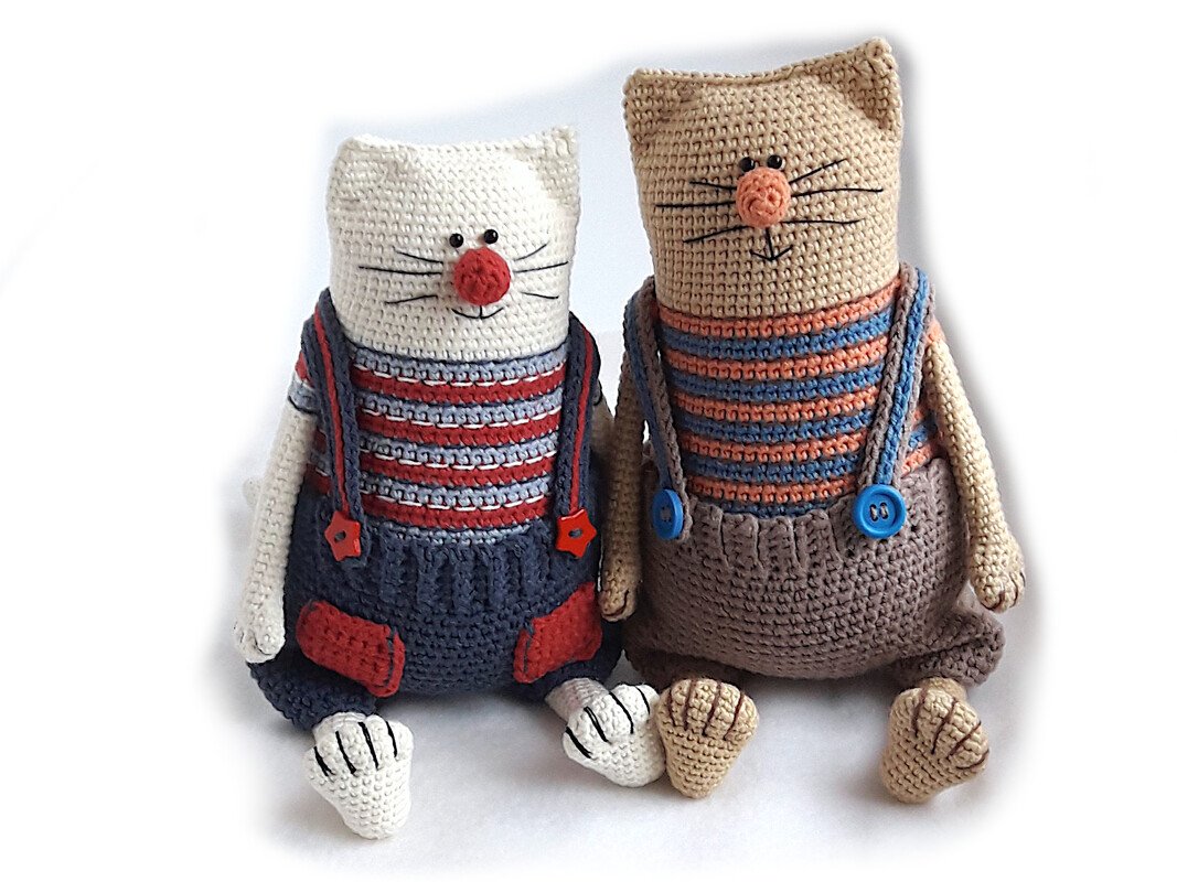 Zwei gehäkelte Amigurumi-Katzen in gestreiften Oberteilen und Latzhosen mit Knopfträgern