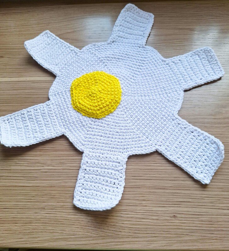 Gehäkelter Topflappen in Form eines Spiegeleis mit gelbem Dotter auf Holzoberfläche.