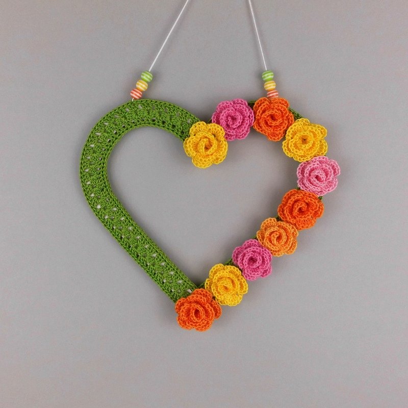 Green crochet heart wreath decorated with yellow, orange and pink crochet rosette flowers hanging by a beaded cord