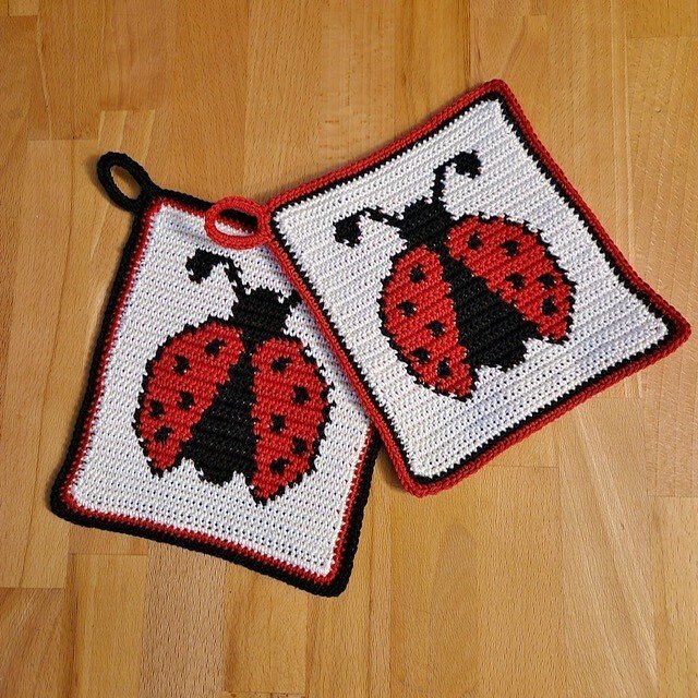 Zwei gehäkelte quadratische Topflappen mit Marienkäfer-Motiv in Rot, Schwarz und Weiß auf Holzuntergrund