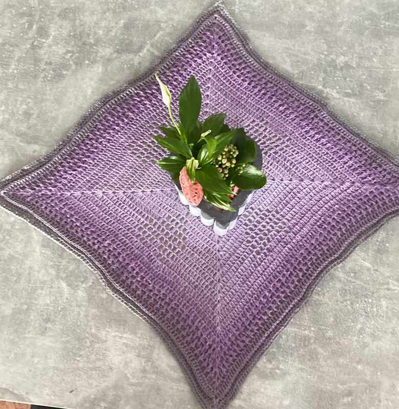Quadratisches lila gehäkeltes Deckchen mit grauem Rand und Topf mit Grünpflanze auf hellgrauem Untergrund