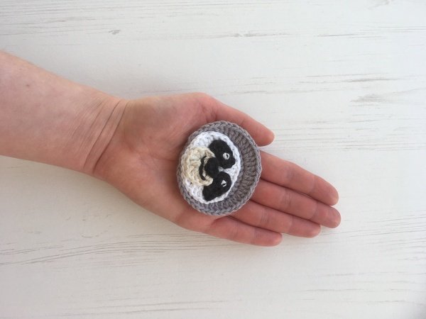 Hand holding small grey and white crocheted sloth face applique