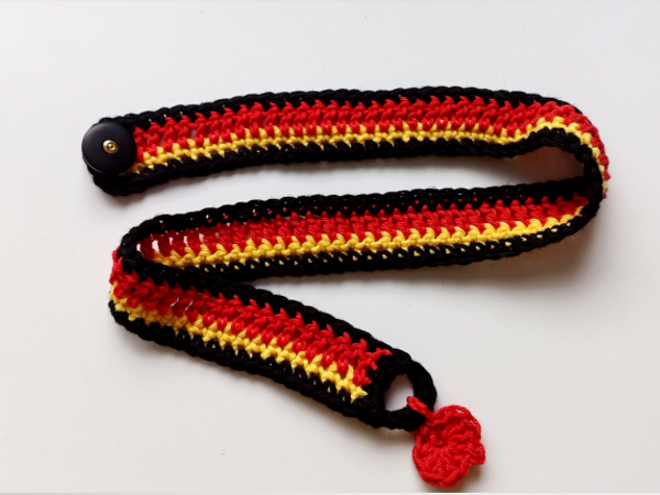 Gehäkelter Riemen in Schwarz, Rot und Gelb mit schwarzem Knopf und kleinem roten Herz auf weißem Hintergrund