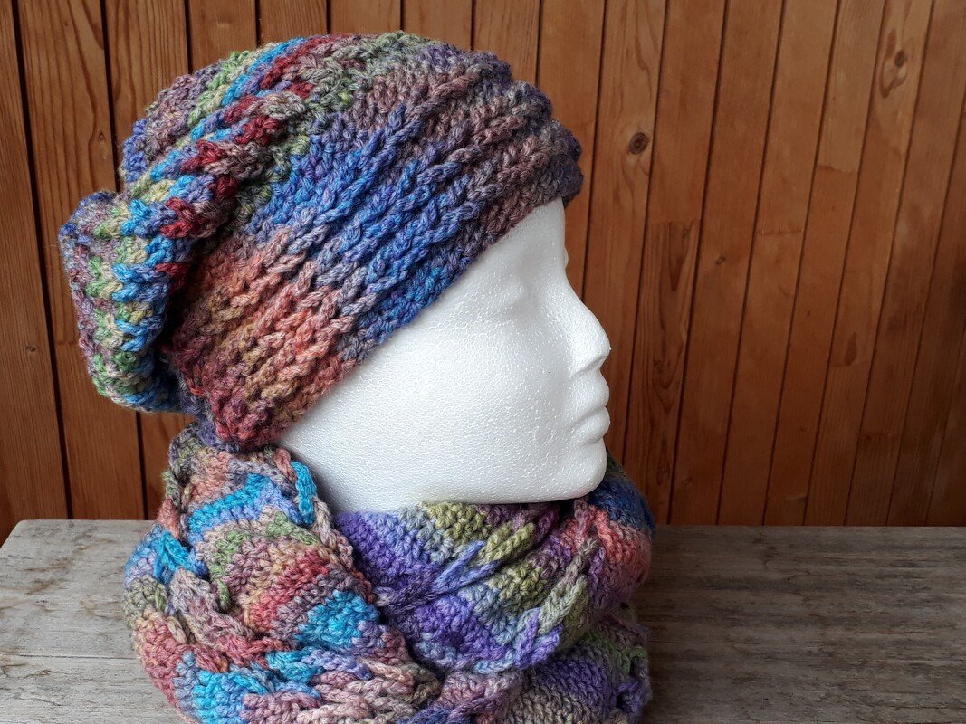 Mehrfarbige gehäkelte Beanie-Mütze und Loop-Schal auf einer weißen Schaufensterpuppe vor einer Holzwand.