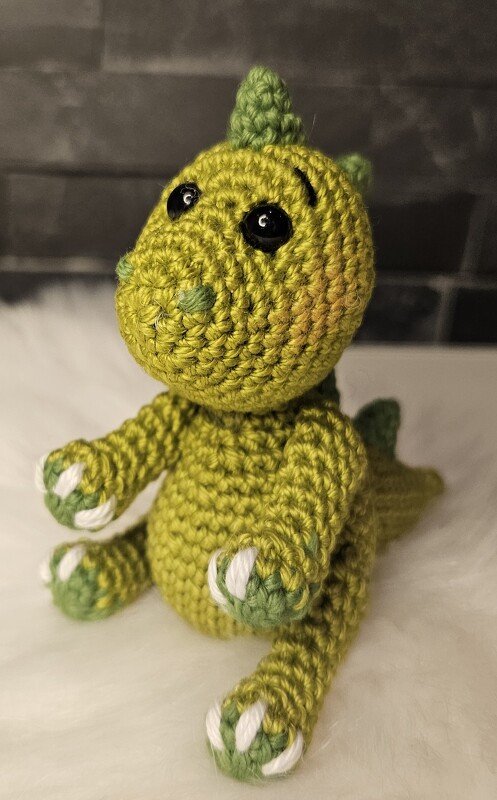 Gehäkelter grüner Amigurumi-Drache mit schwarzen Augen und weißen Krallen, sitzend