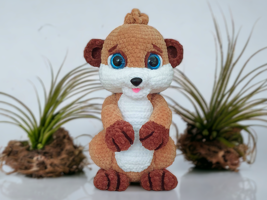 Hand-crocheted brown and white meerkat amigurumi with blue safety eyes and a small pink mouth, standing upright in front of blurred plants.