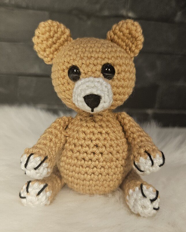 Beiger gehäkelter Teddybär mit weißen Pfoten und schwarzer Nase frontal sitzend