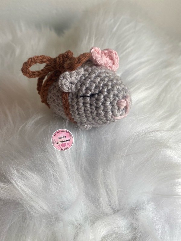Gehäkelte graue Amigurumi-Maus mit rosa Ohren und brauner Schleife auf weißem Flauschstoff
