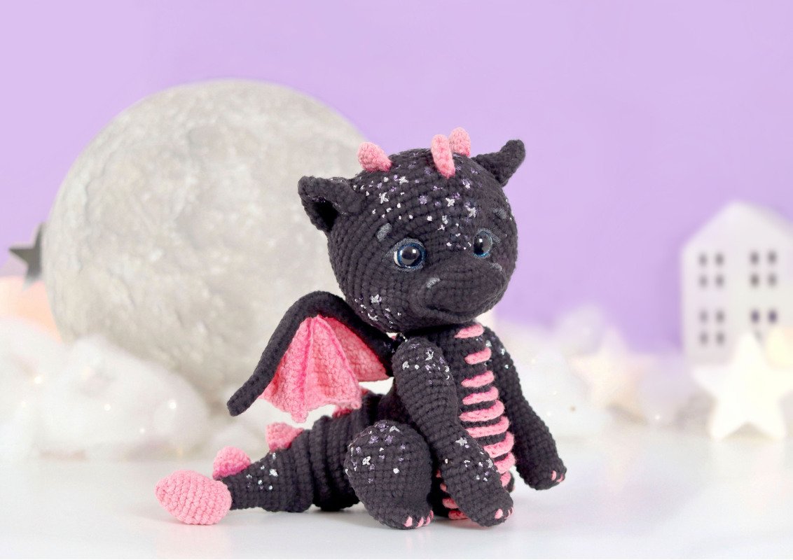 Charcoal crocheted dragon with pink wings and speckled yarn seated in a pastel scene with moon prop