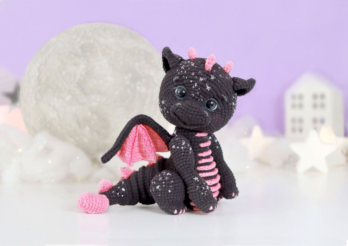 Dunkelgrauer gehäkelter Amigurumi-Drache mit rosa Flügeln und glitzernden Punkten, sitzend vor Mond- und Stern-Dekoration