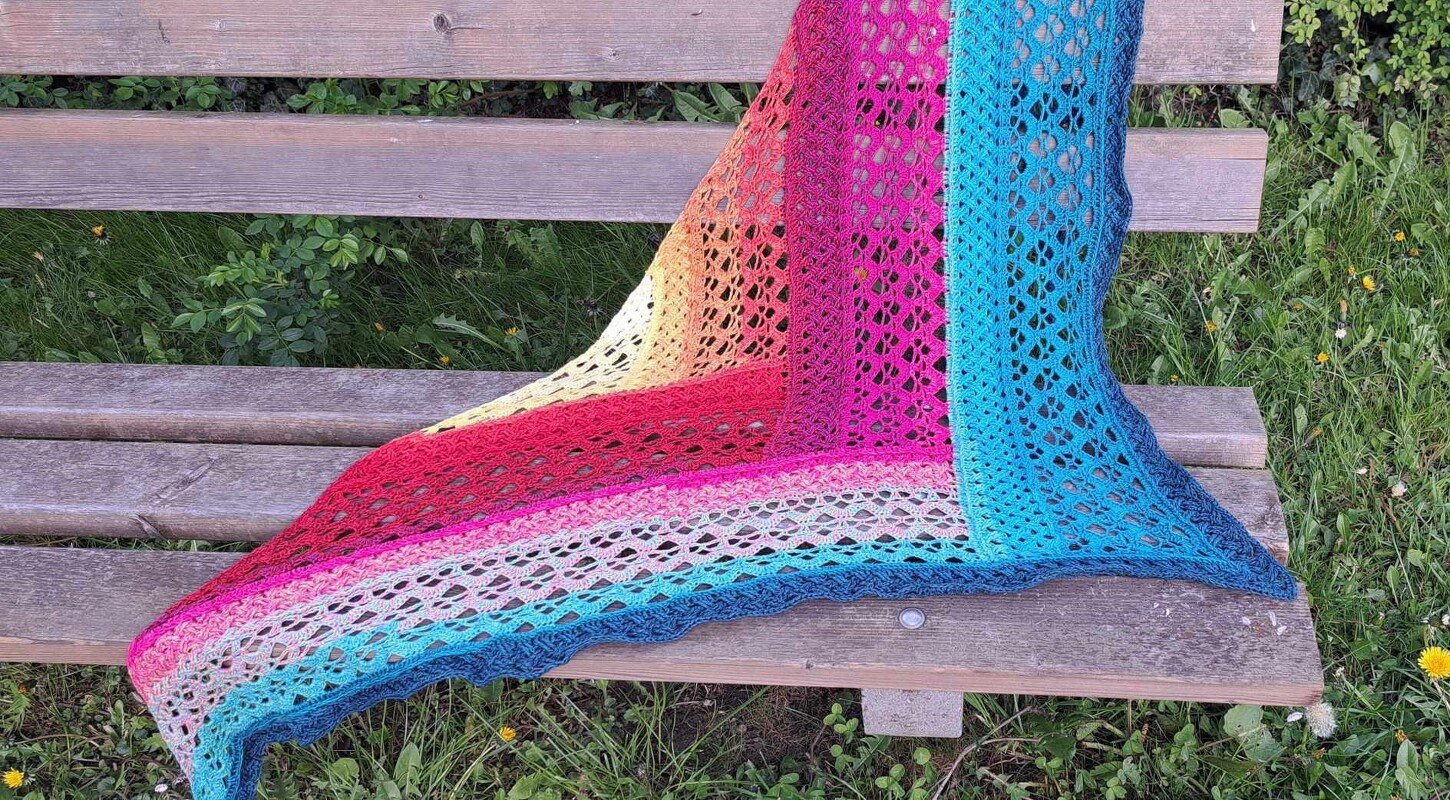 Triangle crochet shawl in rainbow gradient lace draped over a wooden bench on grass