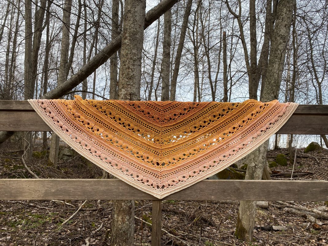 Crochet triangle shawl in golden-to-peach gradient with lace eyelets draped over a wooden fence in a leafless forest