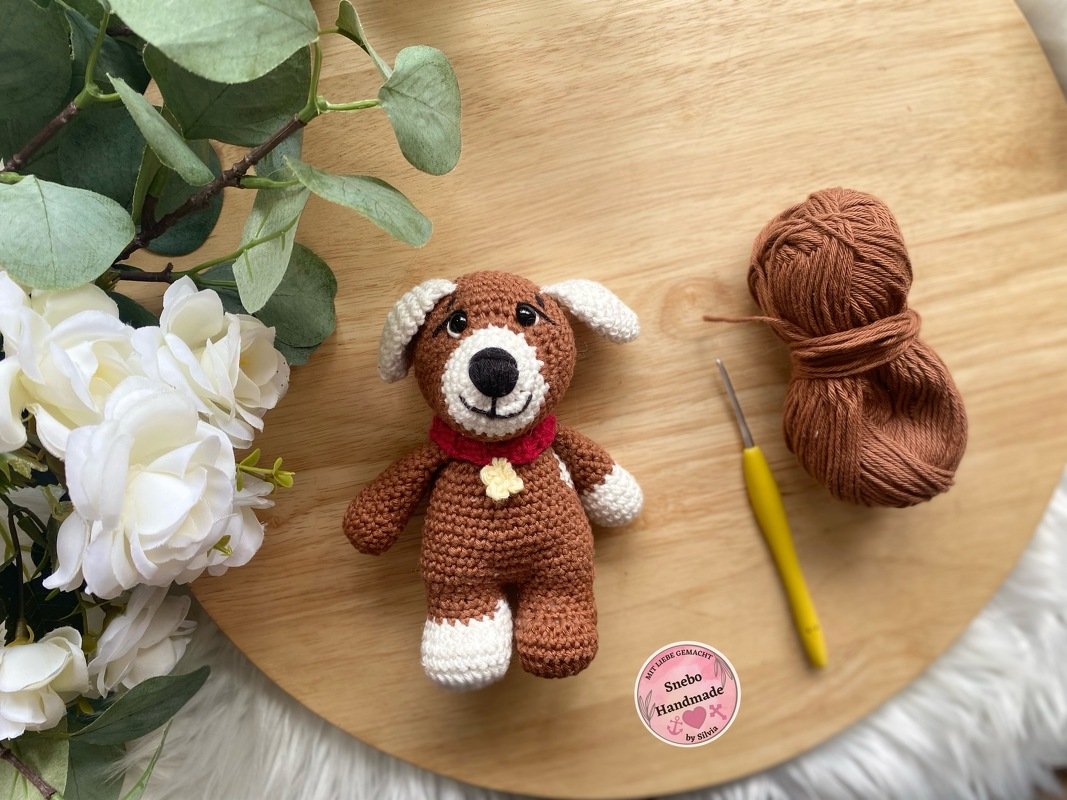 Braun-weißer gehäkelter Amigurumi-Hund mit rotem Halsband, Wollknäuel und gelber Häkelnadel auf Holzplatte