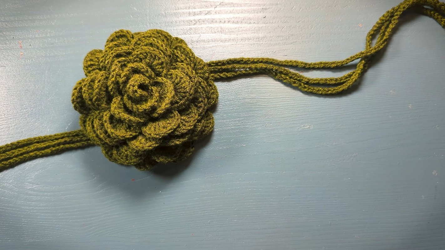 Schmal gehäkeltes Stirnband mit mehrlagiger Blüte und Bindebändern in Oliv auf hellblauem Untergrund