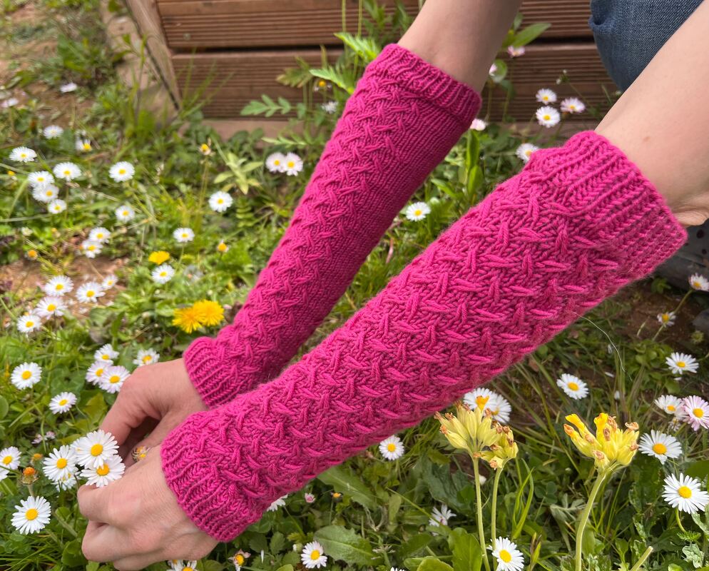 Fingerlose pinke Armstulpen mit Zopfmuster und Daumenöffnung, getragen in einer Blumenwiese