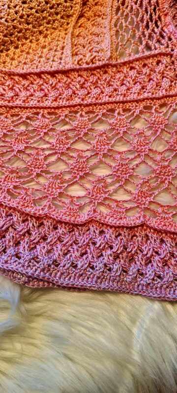 Close-up of a crocheted shawl in peach and pink gradient yarn, showing lace and textured stitch patterns on a soft white surface.