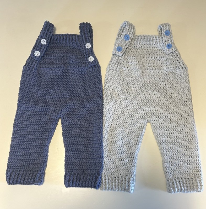 Two crocheted baby overalls, one dark blue with white buttons and one light gray with blue buttons.