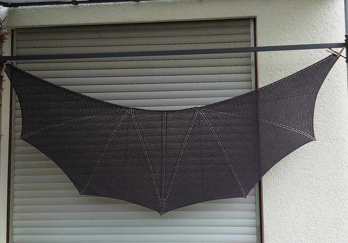 Gehäkeltes dunkles Halbrundtuch mit flügelartigen Bögen, vor einem geschlossenen Rollladen präsentiert