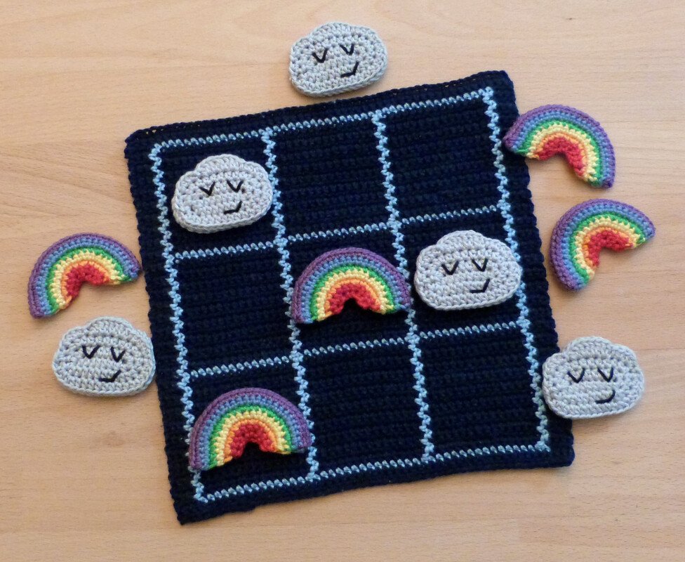 Gehäkeltes Tic-Tac-Toe-Spiel auf hellem Holztisch mit Wolken- und Regenbogen-Spielfiguren, von oben fotografiert.
