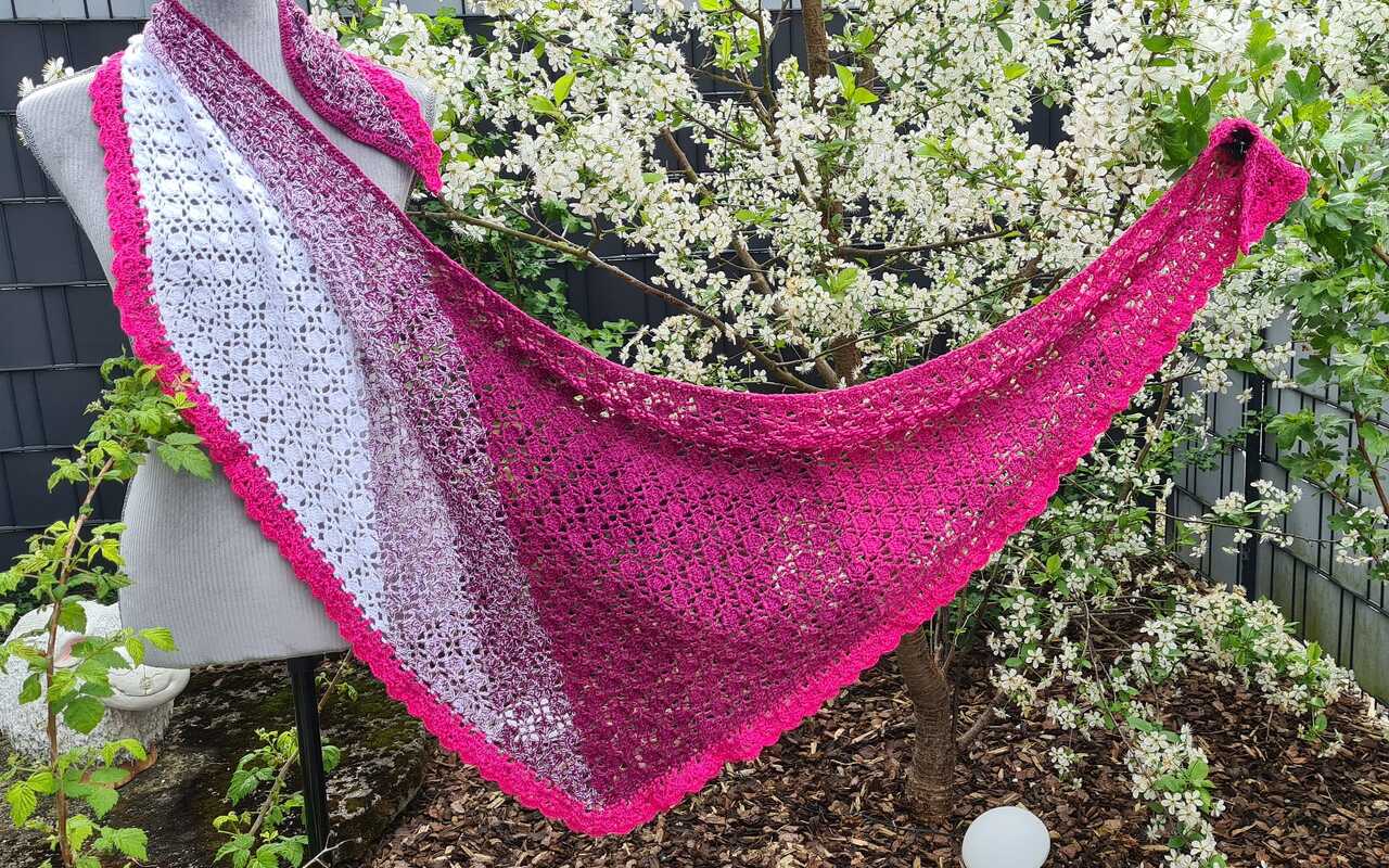 Gehäkeltes Dreieckstuch mit weiß‑zu‑pinkem Farbverlauf, feinem Lochmuster und pinker Muschelkante, an Schaufensterpuppe vor blühendem Strauch drapiert