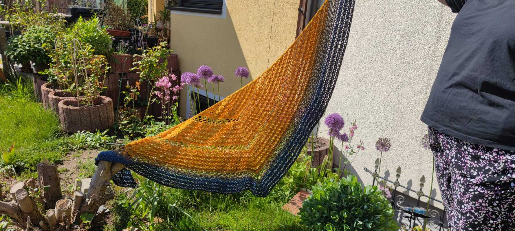 Gehäkeltes Dreieckstuch in Gelb‑Orange zu Blau verlaufend mit Ajourmuster und dunkler Randborte, gehalten im Garten
