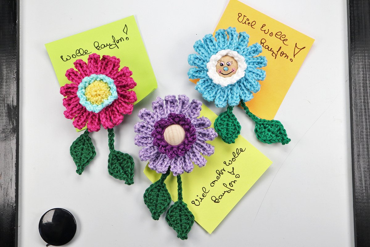 Drei gehäkelte Blumenmagnete in Pink, Lila und Blau auf einer weißen Magnettafel, befestigen Haftnotizen; eine Blume zeigt ein bemaltes Gesicht.
