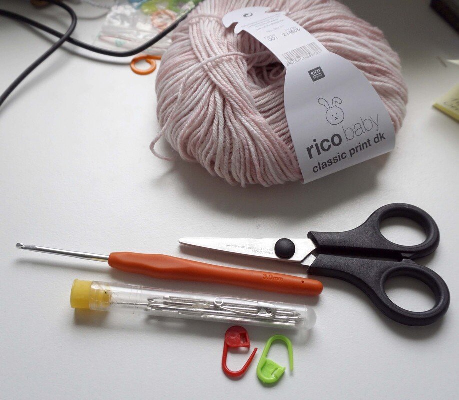 Pale pink DK baby yarn with a 3.0 mm crochet hook, scissors, sewing needles and stitch markers on white background