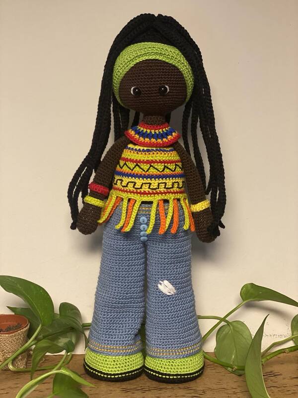 Crocheted amigurumi doll with long black braids, green headband, colorful fringed top and blue flared pants