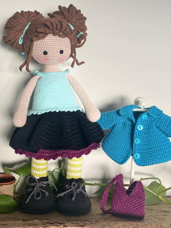 Crocheted amigurumi doll with blue top, black skirt and striped socks next to a blue jacket and purple handbag.