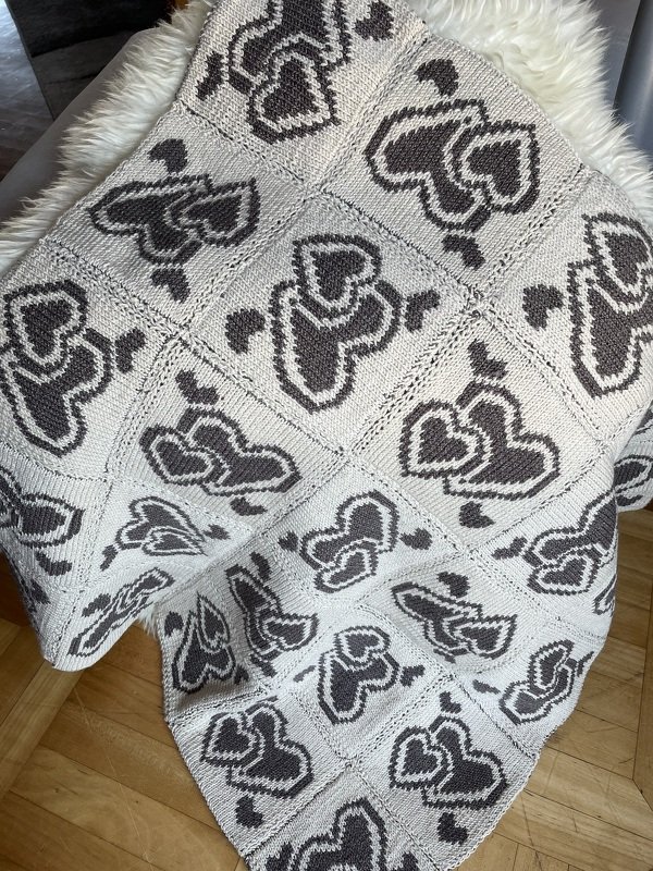 Zweifarbige gestrickte Babydecke aus Quadraten mit Herzmotiven, über einen Stuhl gelegt auf Holzboden