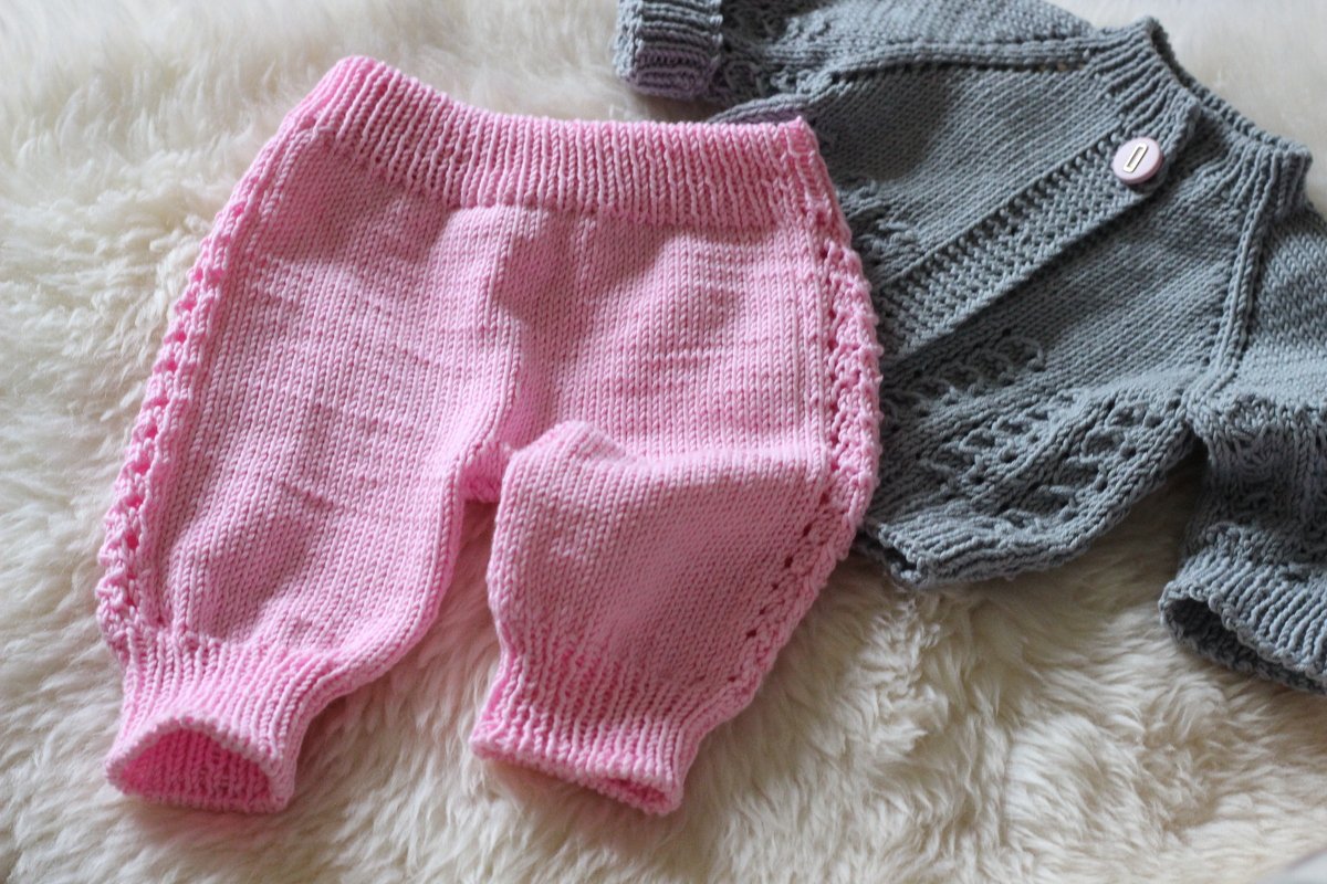 Rosa gestrickte Babyhose mit Lochkante neben grauer Strickjacke auf hellem Fell